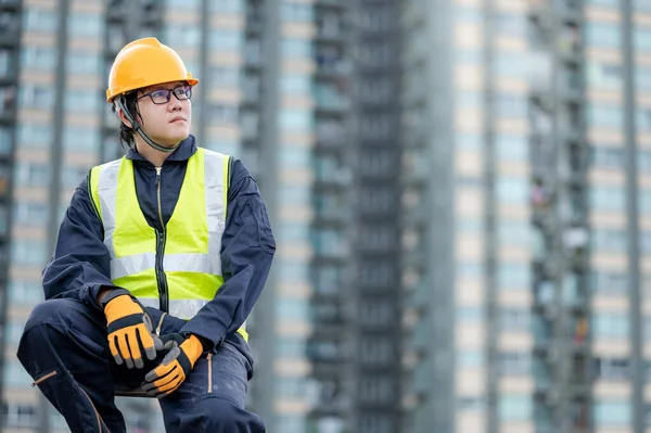 Asian maintenance worker man with safety helmet and green vest sitting on aluminium step ladder at construction site. Civil engineering, Architecture and building service concepts