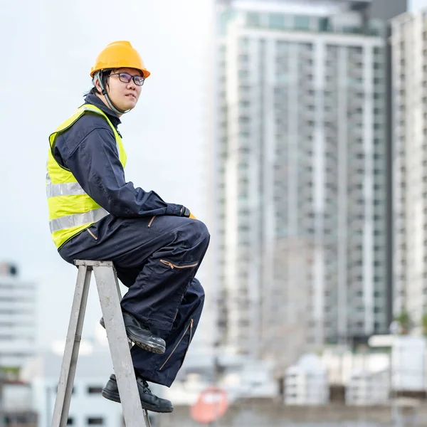 Asian maintenance worker man with safety helmet and green vest sitting on aluminium step ladder at construction site. Civil engineering, Architecture and building service concepts