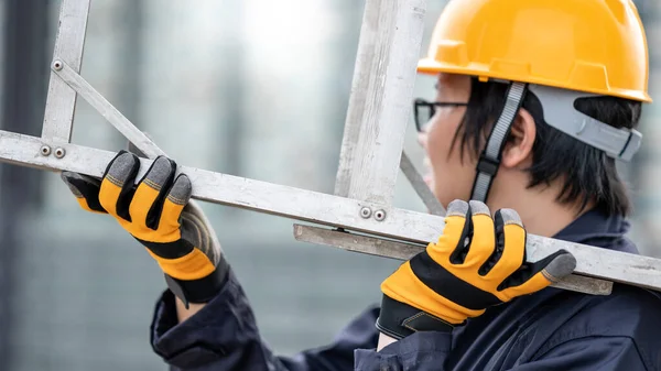 Asian maintenance worker man with protective suit and safety helmet carrying aluminium step ladder at construction site. Civil engineering, Architecture builder and building service concepts