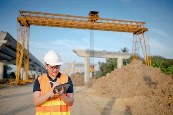 Smart Asian worker man or male civil engineer with protective safety helmet and reflective vest using digital tablet for project planning and checking architectural drawing at construction site.