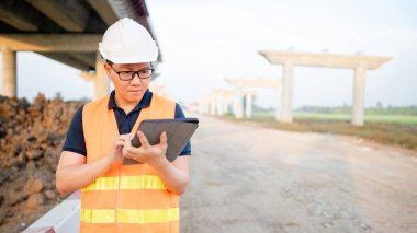 İnşaat alanında proje planlama ve mimari çizimleri için dijital tablet kullanan koruyucu kasklı ve yansıtıcı yelekli akıllı Asyalı işçi ya da erkek inşaat mühendisi..
