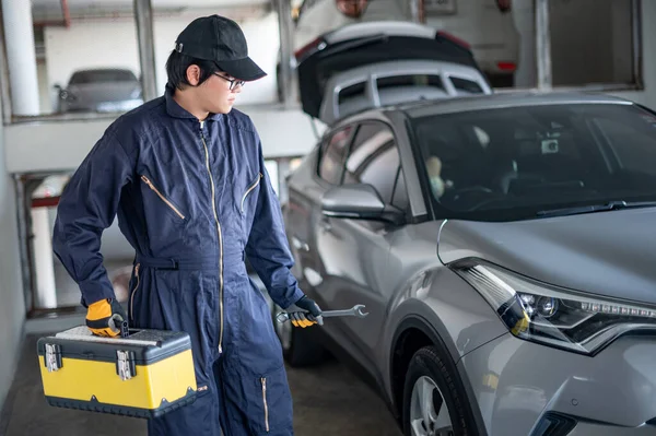 Asian auto mechanic holding tool box and wrench checking car in auto service garage. Mechanical maintenance engineer working in automotive industry. Automobile servicing and repair concept