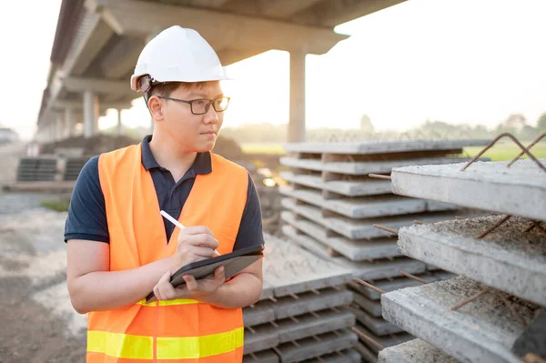 Smart Asian worker man or male civil engineer with protective safety helmet and reflective vest using digital tablet for project planning and checking material at construction site.