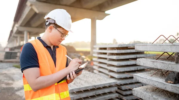 Smart Asian worker man or male civil engineer with protective safety helmet and reflective vest using digital tablet for project planning and checking material at construction site.