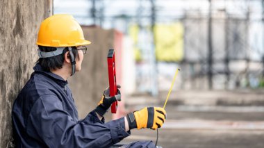 Bakım işçisi ya da Asyalı erkek tamirci koruyucu giysi ve miğfer giymiş ellerinde mezura ve inşaat alanında aletler var. İnşaat mühendisliği projesi için ekipman