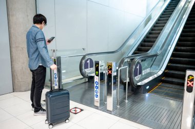 Seyahat sigortası konsepti. Havaalanı terminalindeki yürüyen merdivende bavul taşırken akıllı telefon kullanan Asyalı turist..
