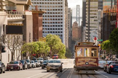 SAN FRANCISCO, CALIFORNIA, ABD - 9 Nisan 2019: Kaliforniya caddesinde ünlü klasik teleferik arka planda San Francisco Körfez Köprüsü ile finans bölgesine doğru bakıyor.