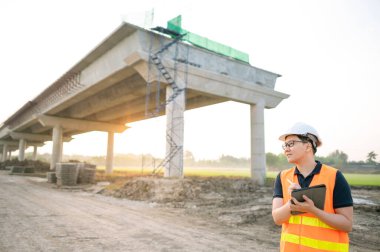 İnşaat alanında proje planlama ve mimari çizimleri için dijital tablet kullanan koruyucu kasklı ve yansıtıcı yelekli akıllı Asyalı işçi ya da erkek inşaat mühendisi..