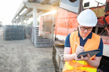 İnşaat alanındaki proje planını kontrol etmek için dijital tablet kullanan kazıcı makinesinin üzerinde koruyucu kaskı ve yansıtıcı yeleği olan akıllı Asyalı işçi veya erkek inşaat mühendisi..