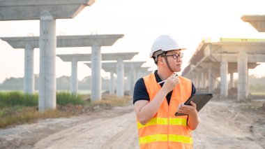 İnşaat alanında proje planlama ve mimari çizimleri için dijital tablet kullanan koruyucu kasklı ve yansıtıcı yelekli akıllı Asyalı işçi ya da erkek inşaat mühendisi..