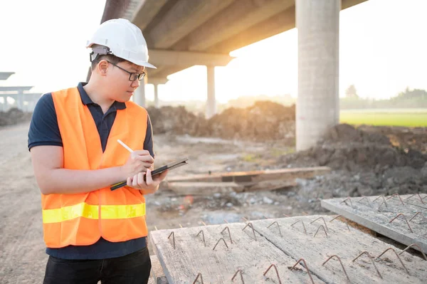 Smart Asian worker man or male civil engineer with protective safety helmet and reflective vest using digital tablet for project planning and checking material at construction site.