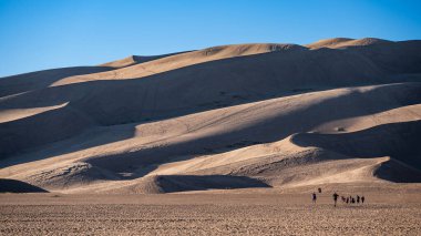 Colorado, ABD 'deki Great Sand Dunes Ulusal Parkı' nda çöl manzarasında yürüyen turistler. Kum tepeciklerinin kıvrımları açık mavi gökyüzüne gölge düşürüyor. Kuzey Amerika 'daki en yüksek kum tepeleri.