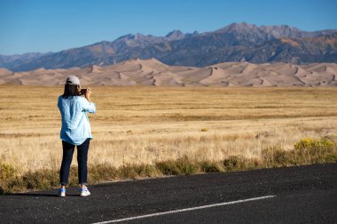 Akıllı telefon kullanan bayan turist, sonbaharda Colorado, ABD 'deki Great Sand Dunes Ulusal Parkı' nda açık mavi gökyüzüne karşı çöl manzarası ve altın çayır fotoğrafı çekiyor..
