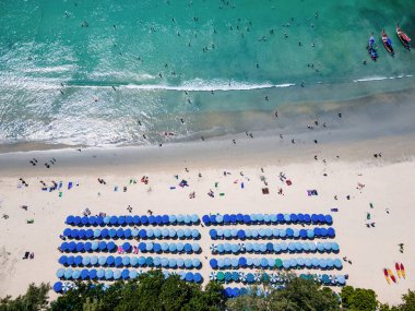 Birçok turistin kumsalda dinlendiği ve Tayland 'ın Phuket kentindeki ünlü bir tatil beldesi olan Kata sahilinde yüzdüğü mavi plaj şemsiyeleri. Andaman Denizi 'nin tropikal kıyılarının havadan görünüşü.