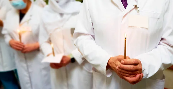 A young man in a medical mask, medical cap and white coat holds a burning candle in front of him. The concept of last hope, faith in a bright future. Focus on the male doctor