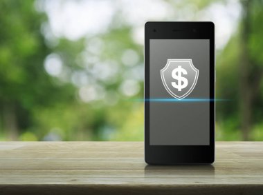 Parktaki bulanık yeşil ağaç, iş parası sigortası ve çevrimiçi korunma kavramının üzerindeki ahşap masa üzerinde modern akıllı cep telefonu ekranında kalkanın düz simgesi olan dolar.