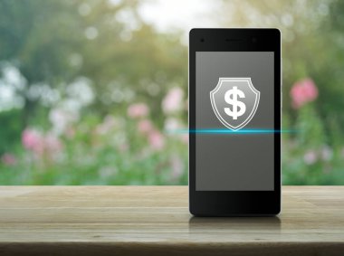 Modern akıllı cep telefonu ekranında paralı simgesi olan dolar, ahşap masa üzerinde bulanık pembe çiçek ve bahçede ağaç, iş parası sigortası ve çevrimiçi koruma kavramı.