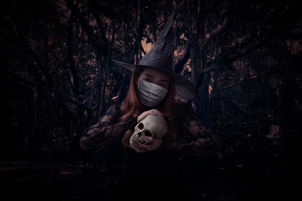 Halloween witch wearing medical face mask holding a skull standing over spooky dark forest with tree, leaves and vine, Halloween and coronavirus or covid-19 concept