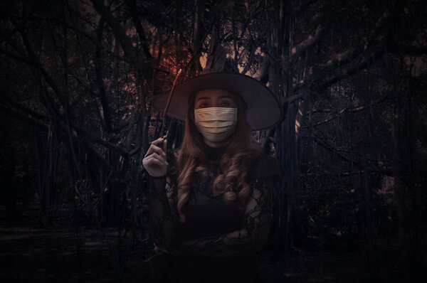Halloween witch wearing medical face mask holding magic wand standing over spooky dark forest with tree, leaves and vine, Halloween and coronavirus or covid-19 concept