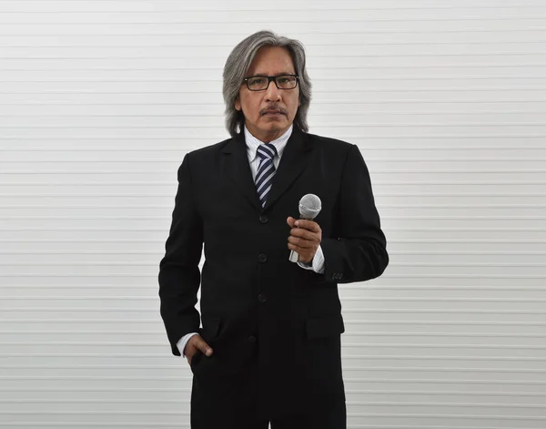 白い壁の背景にマイクを持つ黒のスーツと眼鏡で話す高齢者のアジアのビジネスマンを自信を持って ビジネスプレゼンテーションとセミナーのコンセプト — ストック写真