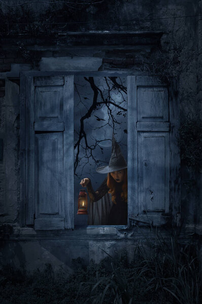 Halloween witch holding ancient lamp standing in old damaged wood window with wall over church, birds, dead tree and spooky cloudy sky, Halloween mystery concept