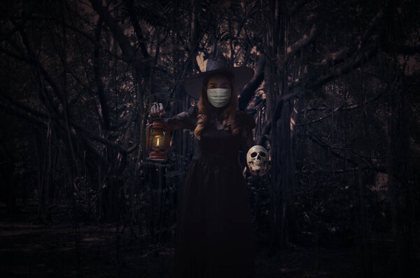 Halloween witch wearing medical face mask holding ancient lamp and skull standing over spooky dark forest with tree, leaves and vine, Halloween and coronavirus or covid-19 concept