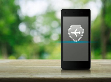 Parktaki bulanık yeşil ağaç, iş seyahati sigortası ve çevrimiçi güvenlik kavramı üzerine modern akıllı cep telefonu ekranında rozeti olan uçak.