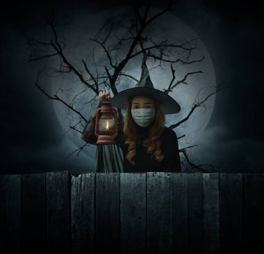 Tıbbi maske takan cadılar bayramı cadısı ölü ağacın, dolunayın ve bulutlu gökyüzünün, Cadılar Bayramı ve Coronavirüs 'ün ya da covid-19 konseptinin üzerinde duran antik lambayı tutuyor.