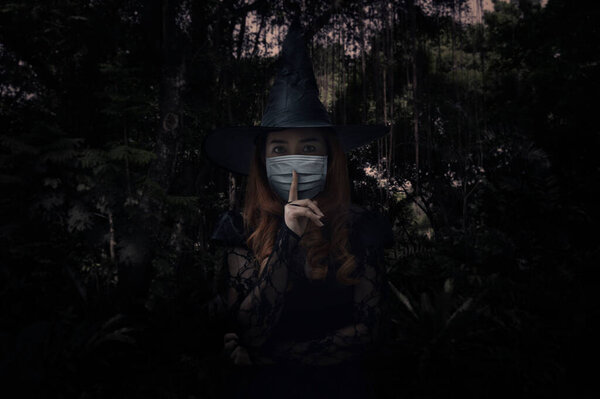 Halloween witch wearing medical face mask showing silence sign with finger over lips standing over dark forest and tree, Halloween and coronavirus or covid-19 concept