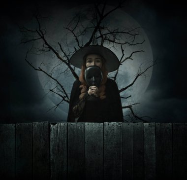 Halloween witch holding black face mask standing over dead tree, full moon and cloudy spooky sky, Halloween mystery concept