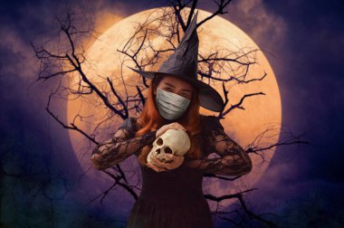 Ölü ağacın, dolunayın ve ürkütücü bulutlu gökyüzünün, Cadılar Bayramı 'nın ve Coronavirüs' ün ya da covid-19 konseptinin üzerinde duran bir kafatası tutan tıbbi maske takan cadılar bayramı cadısı.