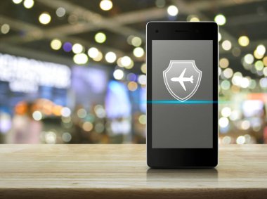 Modern akıllı cep telefonu ekranında silik ışık ve alışveriş merkezinin gölgesi, iş seyahati sigortası ve çevrimiçi güvenlik kavramının üzerindeki ahşap masa üzerinde silikon simgeli uçak.