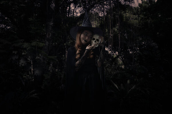 Halloween witch holding a skull standing over dark forest and tree, Halloween mystery concept