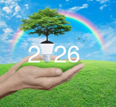 2026 beyaz metin toprakta taze yeşil ağaç ve yeşil çimlerde ampul mavi gökyüzünde, beyaz bulutlar, gökkuşağı ve kuşlar üzerinde mutlu yıllar 2026 yeşil ekoloji ve tasarruf enerji kavramı