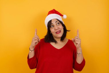 Orta yaşlı, başarılı Arap bir kadının Noel şapkası takıp her iki işaret parmağıyla işaret ederek boş bir fotokopi duvarında şok edici bir şey gösterdiği yatay bir görüntü..