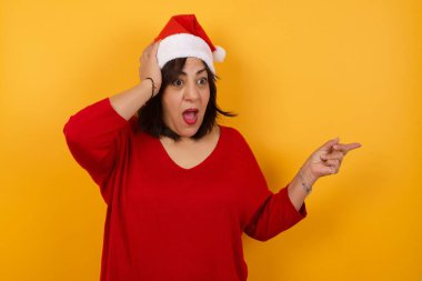Orta yaşlı Arap bir kadının portresi şok olmuş yüz ifadesi ile Noel şapkası takıyor, boş alanda şaşırtıcı bir şey gösteriyor, bir eli kafasında ve işaret parmağıyla işaret ediyor. OMG kavramı.