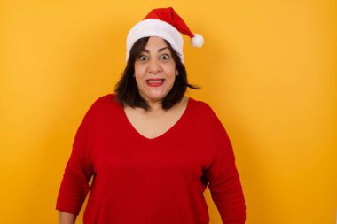 Sarı arka planda duran, geniş beyaz gülüşlü, Noel şapkası takan orta yaşlı Arap bir kadın.