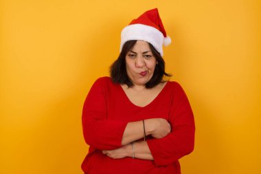 Kızgın, orta yaşlı bir kadının Noel şapkası taktığı ve kollarını kavuşturduğu bir resim. Kameraya bakıyorum..