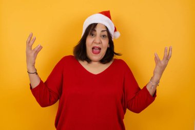 Neşeli Arap orta yaşlı kadın Noel şapkası takıyor, başarıyı kutluyor, sıkıca sarılmış ellerle evet diye bağırıyor.