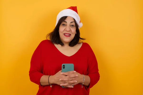 クリスマスの帽子をかぶっているアラブ中年の女性はカメラで注意を向ける スマートフォンの笑顔から気をそらされ 幸せに携帯電話を持って ミラーで自分撮り 友達の新しい服を示す — ストック写真