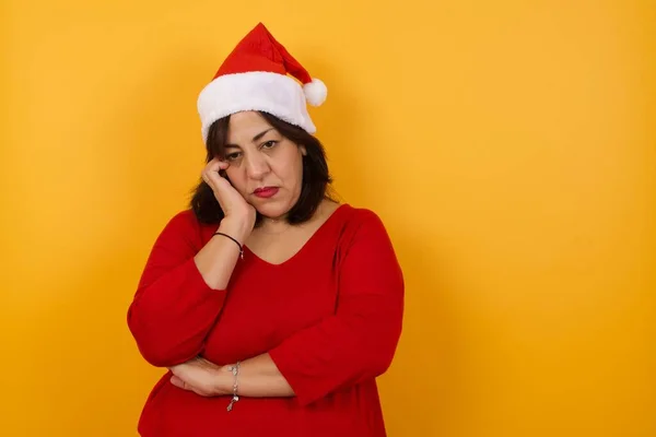 Gloomy arab middle-aged woman wearing Christmas hat  sighing upset stoop look from under forehead lonely unhappy lean palm bored feel regret sadness moody expression
