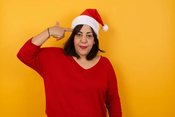 不幸なアラブ中年の女性は 黄色い背景に立っている贈り物でクリスマスの帽子をかぶり 唇を曲げ 銃のジェスチャーを作り すべてに疲れ 手で寺院で撮影し 自殺しようとします 不安コンセプト — ストック写真