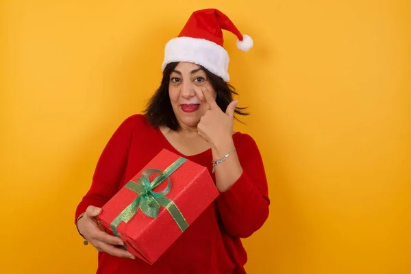 arab middle-aged woman wearing Christmas hat with giftw background, looking, observing, keeping an eye
