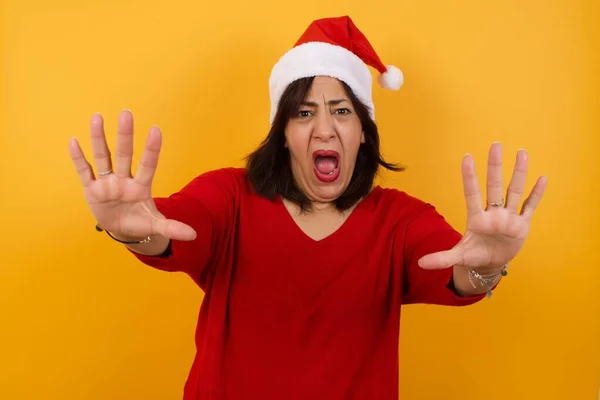 Dissatisfied arab middle-aged woman wearing Christmas hat  frowns face, has disgusting expression, shows tongue, expresses non-compliance, irritated with somebody, rejects do something.