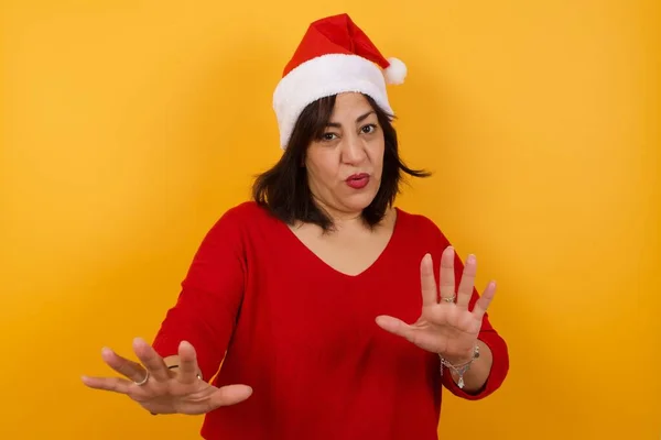 バックグラウンドでクリスマスの帽子をかぶった美しいアラブ中年の女性は恐れを恐れ恐れ 嫌悪感を抱いた表情は両手でジェスチャーをやめます そこにとどまります パニックコンセプト — ストック写真