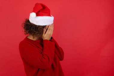 Üzgün, genç, beyaz bir kadın Noel şapkası takıyor. Günlük kıyafetler giyiyor. Kırmızı arka planda duruyor, yüzünü ellerle kaplıyor ve ağlıyor..