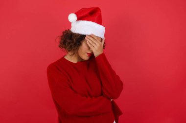 Noel şapkası ve kırmızı süveter giyen güzel bir kadın gülümserken, kırmızı arka planda dikilirken facepalm hareketi yapıyor..