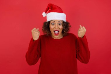 Noel şapkası ve kırmızı süveter giyen, kollarını kaldıran, yumruklarını sıkan, sevinç ve heyecanla bağıran güçlü, kararlı genç bir kadının portresi..