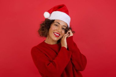 Noel şapkası takan ve güzel bir yüzü olan kırmızı süveter giyen hayalperest genç bir kadın, gözlerini kapatıyor, ellerini yan yana tutuyor, hoş bir şey düşünüyor, kırmızı arka planda poz veriyor..