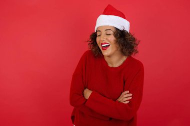 Kıvırcık saçlı, Noel şapkası takan ve kırmızı arka planda kırmızı süveter giyen kıvırcık saçlı genç modelin stüdyo portresi. 